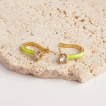 A product photograph showcasing a pair of unique huggie earrings resting on a textured stone surface, finished in a polished gold color and featuring a vibrant apple green enamel accent coat along the back curve, anchored by a prominent round prong-set clear crystal solitaire at the front