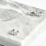 A premium commercial photograph showcasing a pair of modern, open-ended bypass ear cuffs resting on a textured light beige travertine stone. One cuff features a soft pastel pink enamel accent band, while the other features a soft pastel yellow enamel accent band, both beautifully contrasted by a half-track of glittering clear crystals