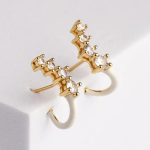 A premium product photograph showcasing a pair of modern post-back stud earrings resting on a white surface. The earrings are finished in polished gold, featuring an ascending curved top bar set with four graduated clear crystals, terminating in a clean, high-gloss white enamel hook