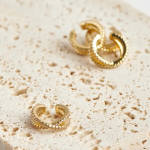 A premium commercial product photograph of a pair of modern post-back stud earrings resting on a textured light cream surface. Each earring is finished in a mirror-polished gold tone, featuring an ascending curved bar set with three graduated, brilliant-cut clear crystals, terminating in a smooth, vibrant sunshine yellow enamel hook