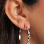 A professional on-ear lifestyle photograph showcasing a large statement hoop earring finished in a polished silver color, lined along its front edge with a continuous vertical row of prong-set clear pear-cut teardrop crystals