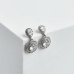 A professional product photograph of a pair of tiered vertical drop earrings finished in high-shine silver color, featuring a teardrop-shaped pear-cut crystal post anchor and a swinging round solitaire crystal drop enclosed in a dramatic double-row micro-pavé crystal halo, resting on a clean white geometric surface