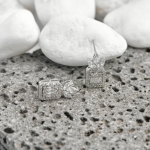 A commercial jewelry product photograph showing a pair of intricate post stud earrings finished in a polished silver color, featuring an upper fan-shaped crown of marquise-cut clear crystals atop a structured rectangular emerald-cut frame encrusted with miniature baguette and micro-crystals, resting on a textured gray basalt rock