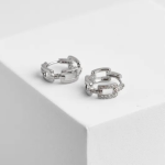 A professional product photograph of a pair of architectural, geometric chain-link huggie hoop earrings finished in a polished silver color, featuring a central rectangular open link encrusted with clear micro-pavé crystals, resting on a clean white geometric backdrop