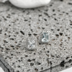 A professional product photograph of a pair of rectangular halo stud earrings finished in a polished silver color, centering a brilliant radiant-cut clear crystal enclosed within a tight, rectangular border of shimmering micro-pavé stones, resting on a textured dark volcanic stone slab