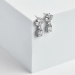 A professional product photograph displayed on a minimalist structural surface, showcasing a pair of mixed-cut geometric drop earrings finished in a polished silver color, featuring a small marquise crystal crown stud that suspends a large, dangling emerald-cut clear crystal encased in a micro-pavé halo