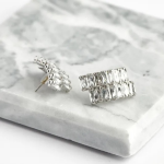 A professional overhead product photograph displayed flat on a white-and-grey veined marble surface, showcasing a pair of geometric shield-shaped stud earrings finished in a polished silver color with a double-row, stacked mosaic layout of clear rectangular baguette-cut crystals