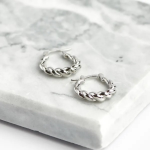 A professional overhead product photograph displayed flat on a white-and-grey veined marble surface, showcasing a pair of sculptural mini huggie hoop earrings finished in a highly polished silver color with a classic, fluidly twisted croissant rope design
