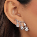 A professional close-up product photograph of a geometric ear crawler earring finished in a polished silver color, featuring a curved lobe bar from which three tiered, emerald-cut clear crystals suspend in graduating lengths, each framed by a matching micro-pavé halo
