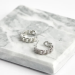 A professional product photograph displayed flat on a veined white marble surface, showcasing a pair of mid-sized hoop earrings finished in a highly polished silver color, designed as a flat cuban link or curb chain layout densely encrusted with clear micro-pavé crystals