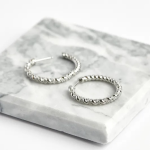 A professional product photograph displayed flat on a veined grey-and-white marble surface, showcasing a pair of delicate, larger-profile hoop earrings finished in a polished silver color, featuring a slender, double-helix intertwined rope design accented by a continuous ribbon of clear micro-pavé crystals