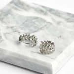 A professional product photograph displayed flat on a veined grey-and-white marble surface, showcasing a pair of delicate botanical huggie hoop earrings finished in a polished silver color, featuring an open-work laurel wreath or olive branch leaf motif encrusted with sparkling clear micro-pavé stones