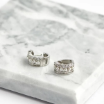 A professional product photograph displayed flat on a veined white marble surface, showcasing a pair of thick, wide huggie hoop earrings finished in a polished silver color, featuring stacked horizontal rows of mini white pearls, round clear crystals, and embedded baguette-cut stones