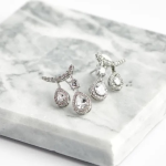 A professional product photograph of a pair of multi-drop jacket earrings finished in a polished silver color, featuring a micro-pavé crescent post anchor from which two staggered, halo-encrusted pear-cut clear crystal drops suspend, resting gracefully on a textured white quartz slab