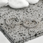 A commercial jewelry product photograph showing a pair of intricate, multi-row open ear cuffs finished in a polished silver color, featuring one delicate scalloped crystal strand and a parallel chevron-woven micro-pavé band, resting on a textured gray basalt slab