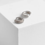 A professional product photograph displayed on a minimalist structural surface, showcasing a pair of chunky, thick huggie hoop earrings finished in a highly polished silver color, featuring a rounded, domed profile densely encrusted with a multi-row carpet setting of clear micro-pavé crystals