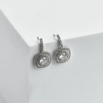 A professional product photograph displayed on a minimalist white architectural surface, showcasing a pair of geometric drop earrings finished in a polished silver color, featuring a small crystal-encrusted linear bar stud that suspends a rounded square cushion-cut central stone framed by two concentric tracks of clear micro-pavé halos