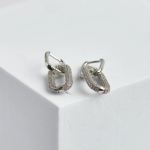 A professional product photograph displayed on a modern, architectural geometric surface, showcasing a pair of interlocking drop earrings finished in a polished silver color, featuring a smooth mini huggie hoop that suspends a dangling, elongated rectangular hardware link densely encrusted with clear micro-pavé stones