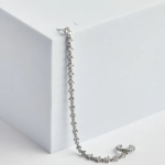 A professional lifestyle product photograph displayed flat on a white-and-grey veined marble surface, showcasing an asymmetric statement earring finished in a polished silver color, featuring a delicate clear solitaire stud connected via a fluid, draping tennis-style crystal chain to an adjustable, micro-pavé ear cuff