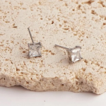 A professional product photograph of a pair of minimalist princess-cut solitaire stud earrings finished in a polished silver color, featuring a sharp, square-cut clear crystal held securely in a clean four-prong setting, resting on a textured cream travertine slab
