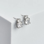 A professional product photograph displayed on a minimalist structural surface, showcasing a pair of elegant statement stud earrings finished in a polished silver color, featuring a small round brilliant clear stone vertically stacked on top of a larger, multi-faceted cushion-cut clear gemstone