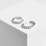 A professional product photograph of a pair of dainty mini huggie hoop earrings finished in a bright silver color, featuring a continuous front-facing track of small, uniform, high-luster white micro pearl spheres, resting on a white geometric cube