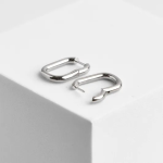 A professional product photograph of a pair of minimalist, elongated rectangular carabiner-style huggie hoop earrings finished in a high-shine, smooth polished silver color, resting flat on a white geometric cube