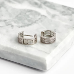 A professional product photograph of a pair of wide, openwork huggie hoop earrings finished in a high-shine silver color, featuring two parallel outer tracks lined with glittering micro-pavé clear crystals flanking an airy, open lattice center lane, resting on a marbled white quartz slab