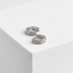 A professional product photograph of a pair of mid-width huggie hoop earrings finished in a polished silver color, featuring two parallel rows of shimmering clear micro-pavé crystals separated by a clean, polished central channel, resting on a white geometric cube