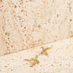 A pair of delicate, gold-tone stud earrings shaped like minimalist crosses, intricately accented with tiny pavé crystals, resting on a textured stone surface