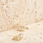 A pair of delicate gold-tone huggie earrings featuring two distinct crystal shapes—a rectangular baguette and a brilliant round solitaire—resting on a textured stone background