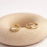 A pair of elegant, gold-tone huggie hoop earrings featuring a twisted rope texture accented with a subtle line of shimmering clear crystals, displayed on a textured beige ceramic surface