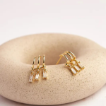 A commercial jewelry flat-lay photograph showcasing a pair of polished gold-colored multi-band claw stud earrings resting on a porous stone display plate, each grounding three independently suspended, vertical rectangular baguette-cut clear crystals