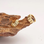 A commercial jewelry flat-lay photograph showing a pair of wide, chunky gold-colored shield-style huggie earrings detailed with alternating vertical sections of polished metal panels and glittering clear micro-pavé crystals, resting on a rough, dark gray volcanic stone