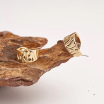 A commercial jewelry photograph showing a pair of thick, chunky gold-colored crescent hoop earrings featuring a heavily textured, organic crumpled foil surface, resting on a rough, dark gray volcanic rock formation