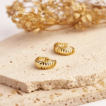 A commercial jewelry flat-lay photograph showing a pair of chunky, polished gold-colored huggie earrings featuring a hollow cage-like design with evenly spaced vertical rib cutouts, resting on a textured stone display piece