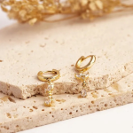 A jewelry flat-lay photograph showcasing a pair of polished gold-colored huggie hoop earrings resting on a porous stone plate, each holding an intricate, dangling marquise-crystal floral cluster that terminates in a free-moving pear-cut teardrop stone