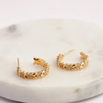 A commercial jewelry flat-lay photograph showing a pair of wide, open-crescent gold-colored stud earrings sculpted into an interlocking botanical laurel leaf or chevron wheat grain pattern, with alternating leaf segments encrusted in sparkling micro-pavé crystals, resting on a white marble platform