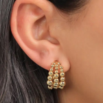 A close-up lifestyle photograph showing a model wearing a chunky, multi-layered gold-colored huggie hoop earring composed of three parallel rows of polished, seamless metallic spheres that taper toward the earlobe