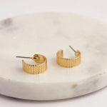 A commercial jewelry flat-lay photograph showing a pair of wide, flat gold-colored open crescent C-hoop earrings featuring a deeply carved vertical fluted column pattern, resting on a smooth white marble platform