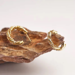 A commercial jewelry photograph showing a pair of open crescent hoop earrings resting on a raw volcanic stone, featuring an intertwined dual-strand design that wraps a smooth, polished gold ribbon with a line of glittering clear pavé crystals
