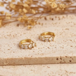 A pair of gold-tone huggie hoop earrings featuring five round, clear crystals arranged in a graduated, light-catching arc along the front