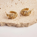 A commercial jewelry flat-lay photograph showing a pair of thick, textured gold-colored crescent hoop earrings featuring an overlapping serpent-scale layout encrusted with alternating rows of vertical crystal baguettes and shimmering micro-pavé, resting on a stone display slab