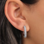 A pair of small, elegant silver-tone huggie hoop earrings fully encrusted with shimmering, round-cut clear pavé crystals, shown being worn on an earlobe