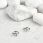 A pair of petite, silver-tone huggie hoop earrings featuring a central pear-shaped crystal surrounded by a delicate pavé halo, displayed on a smooth white stone surface