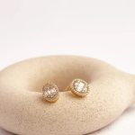 A pair of elegant, gold-tone stud earrings featuring a central baguette-cut crystal framed by a shimmering micro-pavé halo, displayed on a textured stone surface
