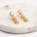 A pair of sophisticated gold-tone drop earrings featuring three linked rectangular segments, each centered with a baguette-cut crystal and framed by a halo of pavé stones, resting on a white marble surface