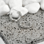 Elegant silver-tone huggie hoop earrings featuring a sophisticated, intertwining twisted design accented with delicate pavé crystals, displayed on a textured stone surface