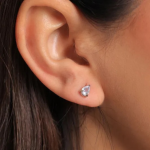 A close-up photograph of a model's ear showing a tiny, polished silver-colored post earring holding a single, brilliant pear-cut clear crystal solitaire