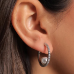 A close-up lifestyle photograph of a model wearing a modern, open-back silver-colored hoop earring encrusted with clear micro-pavé crystals, beautifully curving around the earlobe to suspend a round, luminous white pearl inside the hoop's orbit